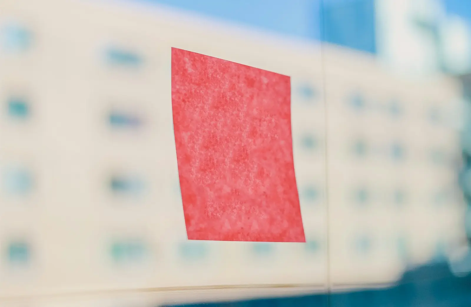 A blank red sticky note stuck to a window, with a pale building blurred in the background