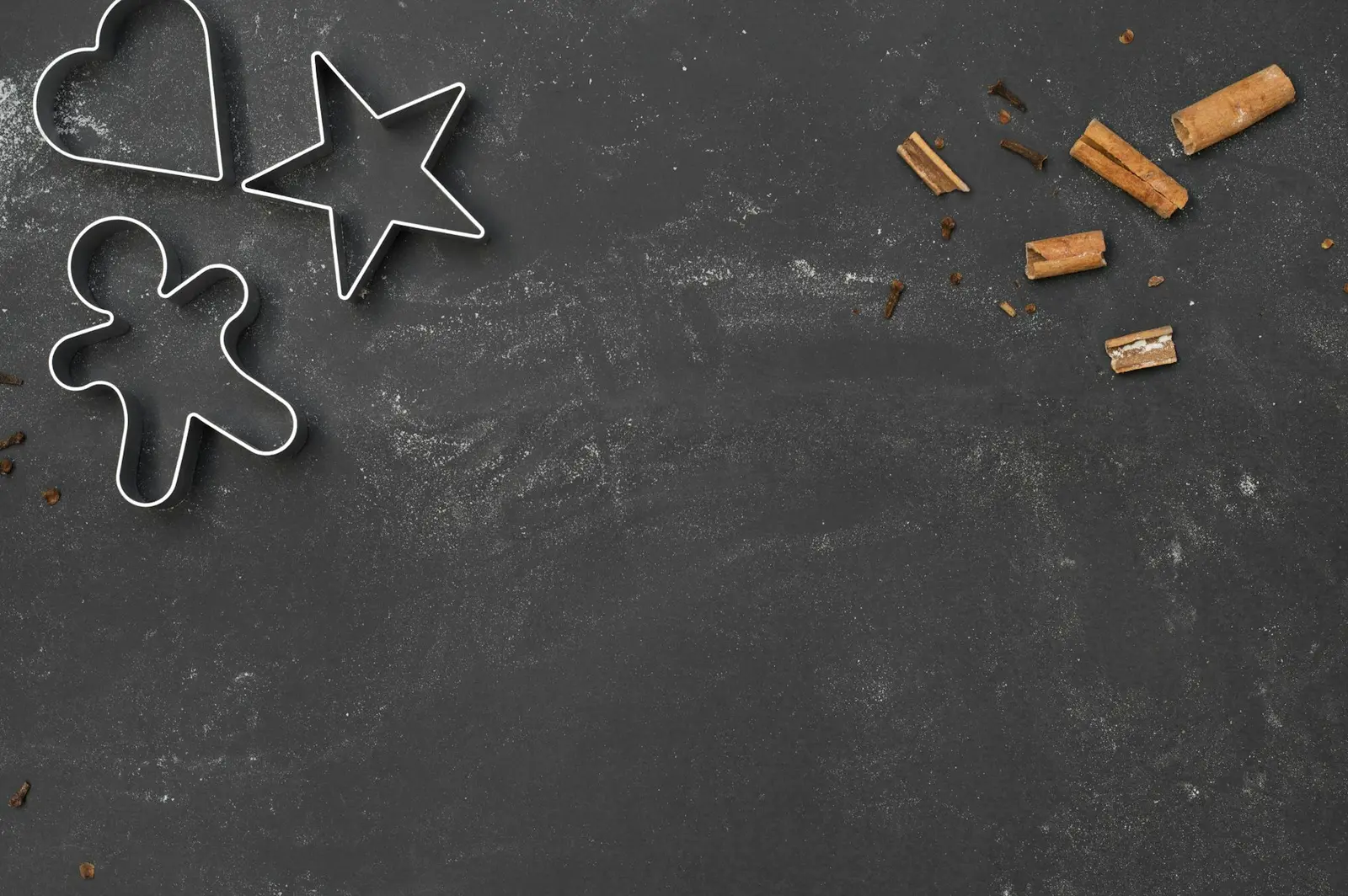 Three metal cookie cutters shaped like a heart, a star and a gingerbread figure on a dark floury surface, with cinnamon sticks to one side