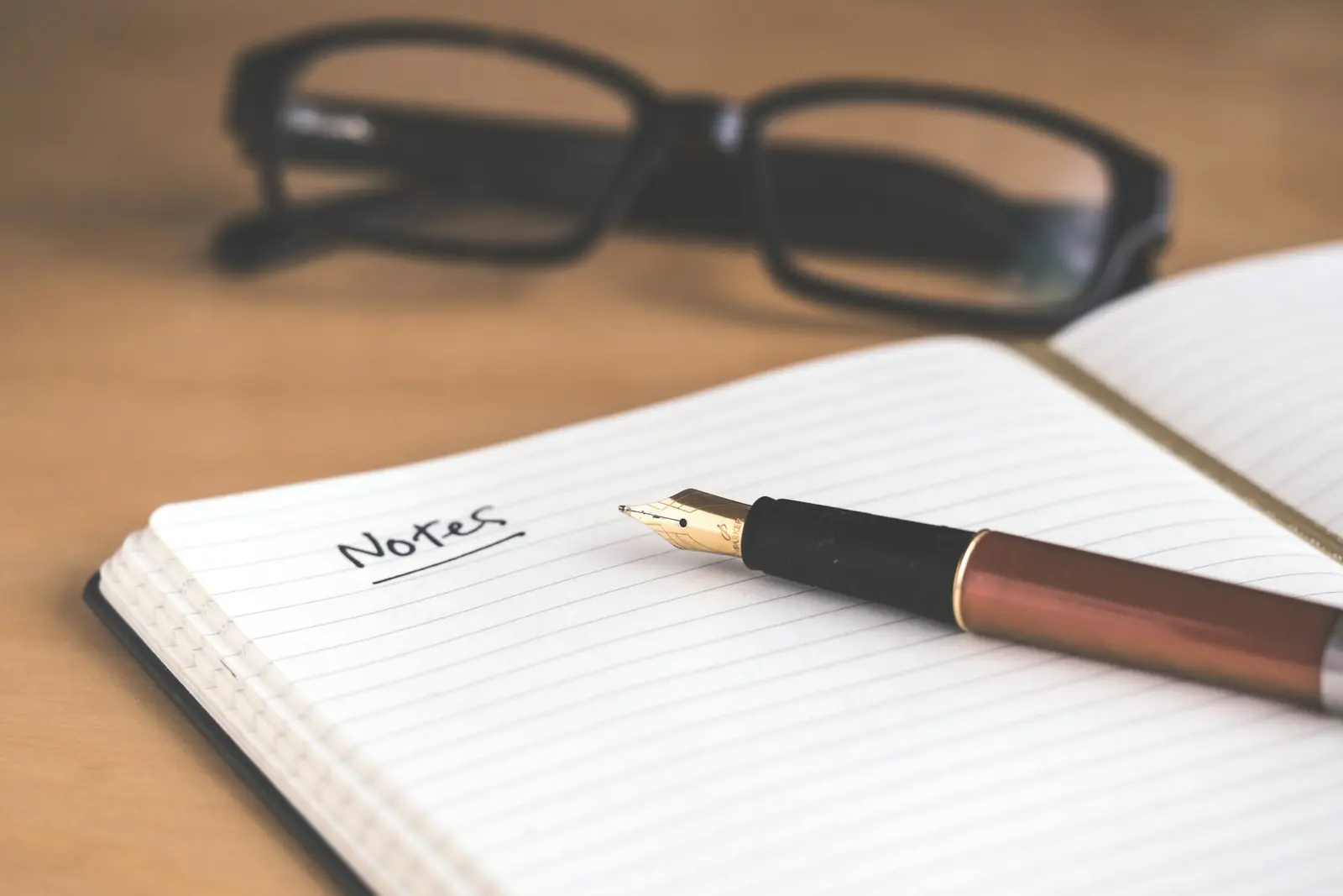 A fountain pen lying across an open ruled notebook with the word Notes handwritten at the top and reading glasses blurred behind it