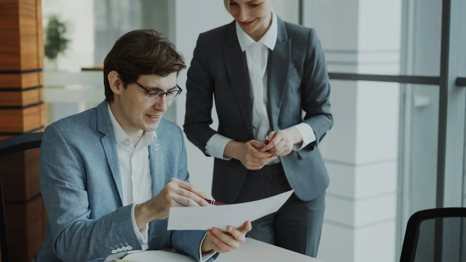 A man in a grey-blue blazer and glasses sitting at a table and holding a sheet of paper with a pen, while a woman in a dark suit stands beside him looking down at it, with no text on the sheet visible