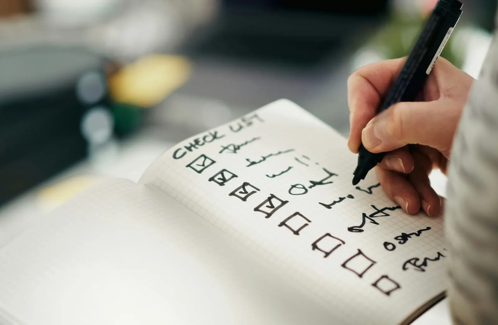 A hand with a black marker ticking boxes on a handwritten checklist in a squared notebook