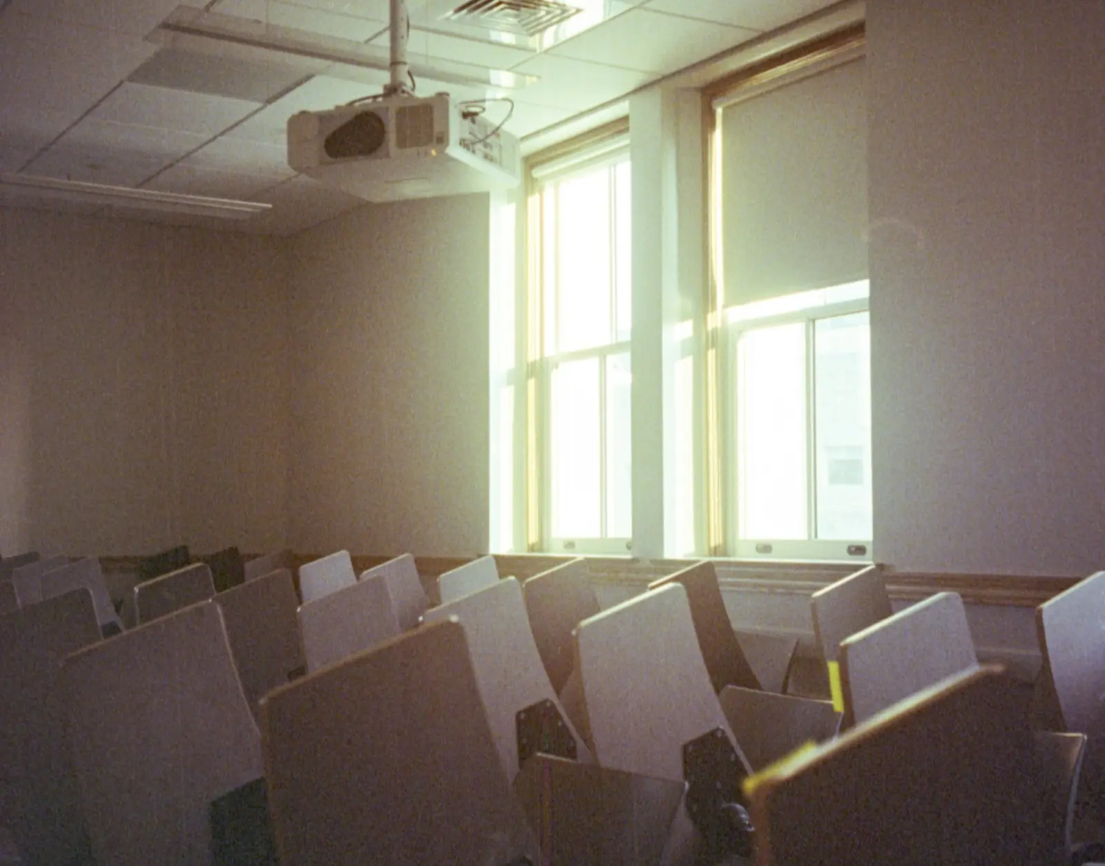 An empty lecture room with rows of grey fold-down seats, a ceiling projector and tall windows letting in daylight