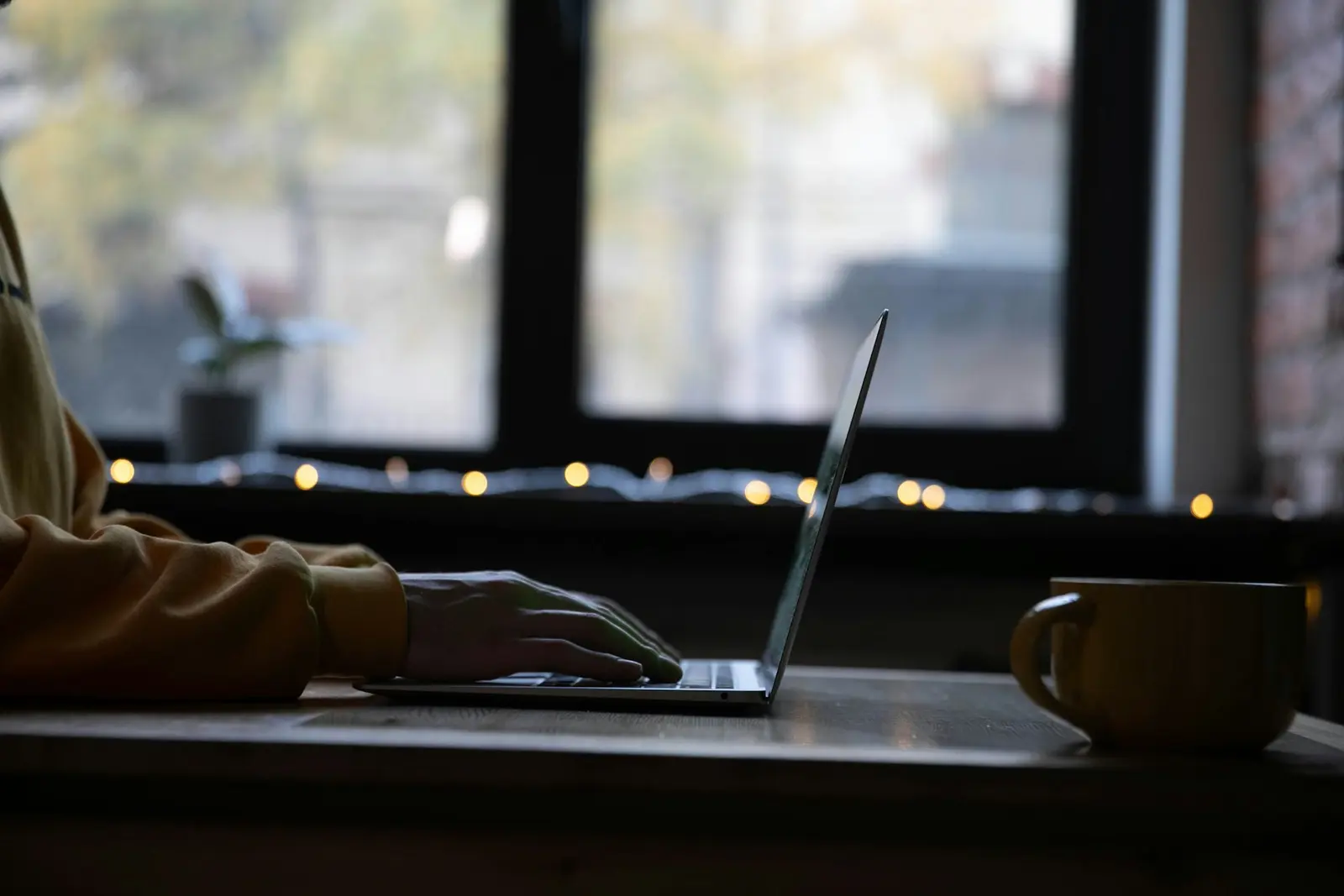 A person's hand on the keyboard of an open laptop on a dark desk beside a mug, in front of a window with string lights, seen in silhouette