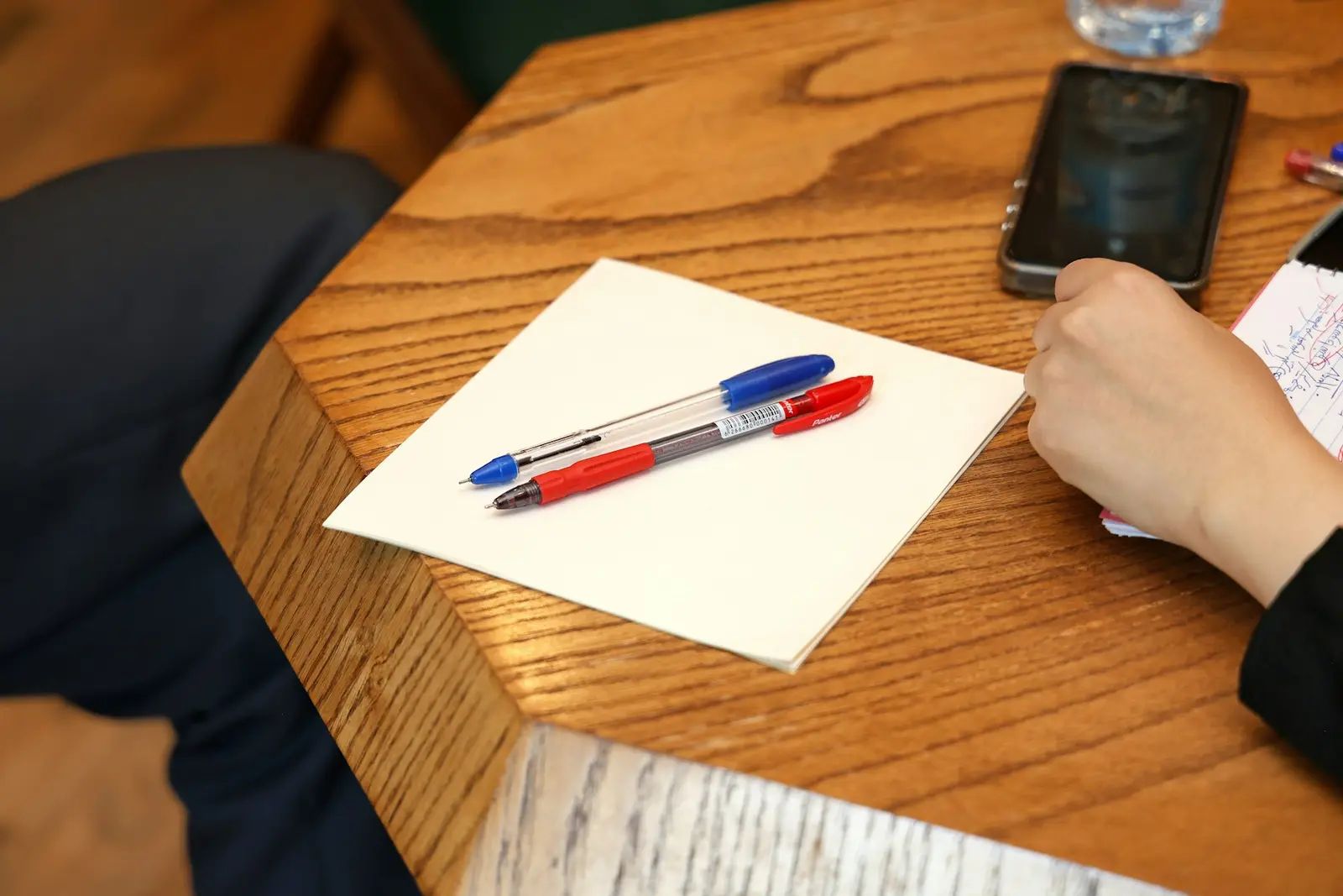 A red pen and a blue pen lying on a white notepad on a wooden table, with a hand and a phone at the edge of the frame