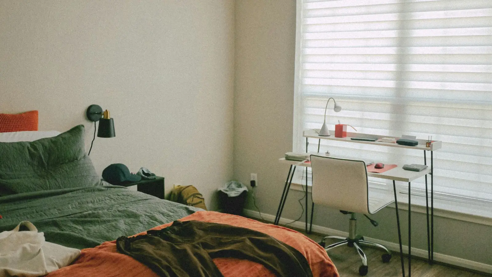 A small bedroom with a green and orange bed, a black wall lamp, and a white desk and chair under a window with pale blinds