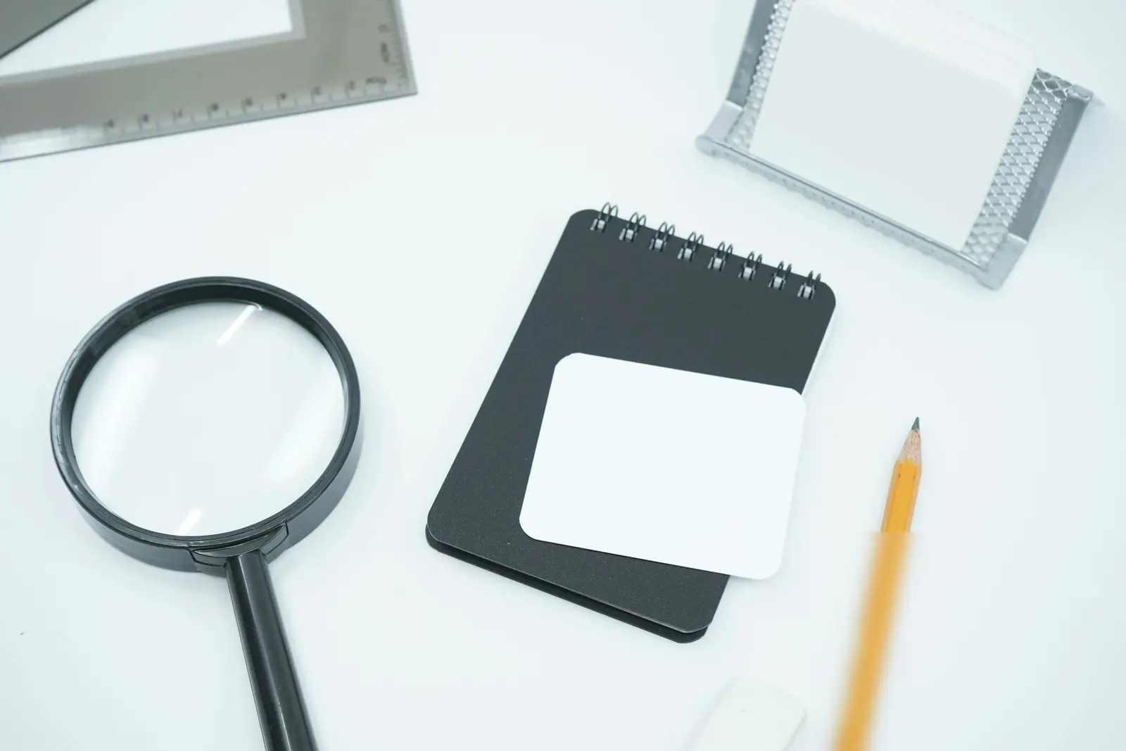 A black magnifying glass next to a small black spiral notepad with a blank white label, a pencil and a metal ruler on a white desk