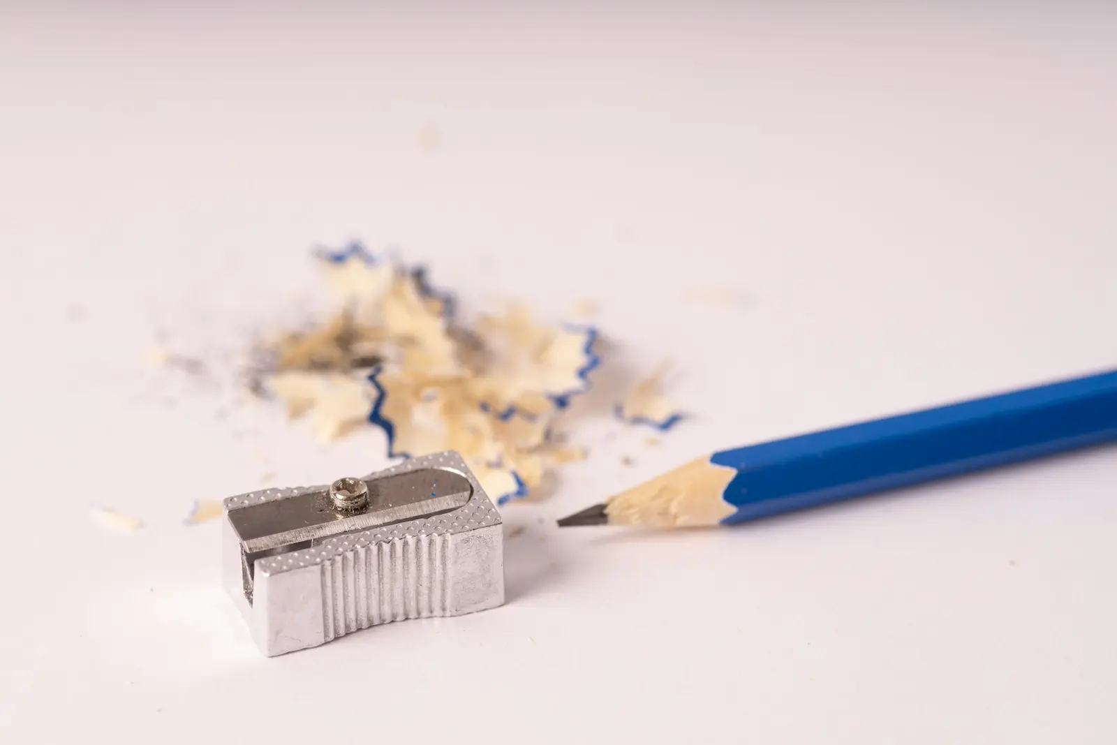 A blue pencil with a sharpened tip lying beside a metal pencil sharpener and a small pile of wood shavings on a white surface